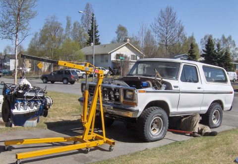1978 Ford Bronco - Mechanic works on a 1978 Ford Bronco, with a separate engine being lifted by a hoist.
