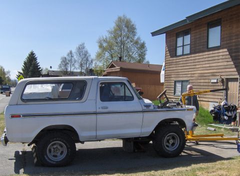 1978 Ford Bronco - A white 1978 Ford Bronco is parked outside a wooden house while an engine is being removed with a yellow engine hoist.