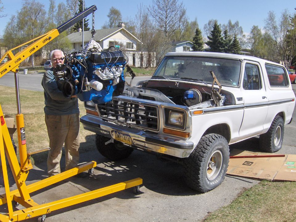 White 1978 Ford Bronco with hood open, engine being lifted out by a yellow engine hoist