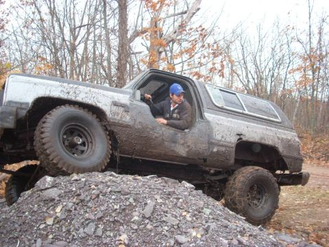 1988 Chevrolet Blazer - A driver navigates a muddy, lifted SUV over a rocky incline in a wooded area.