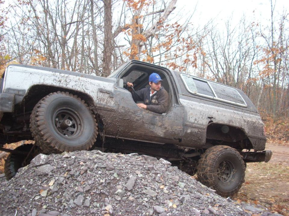 Muddy SUV climbing a pile of rocks with a driver inside. The vehicle appears to be a Chevrolet K5 Blazer from the late 1980s