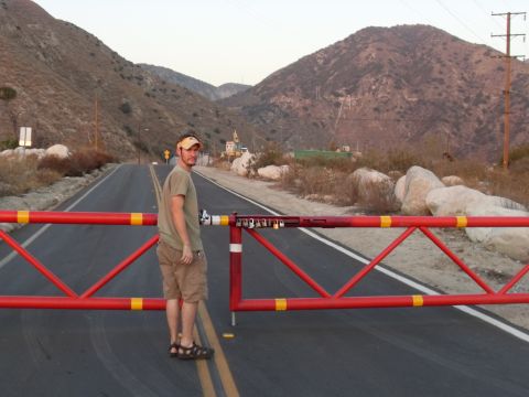 1990 Ford Bronco - Man stands by a red road barrier on a paved road in a mountainous area. Supermotors logo in lower right.