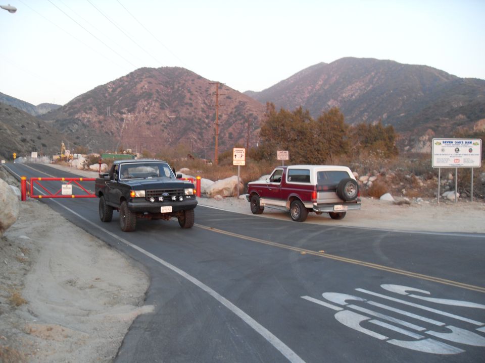 Red and white Ford Bronco and black Ford F-150 truck parked on a road near a gate and mountains
