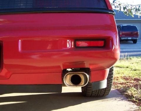 1988 Pontiac Fiero - The rear of a red 1988 Pontiac Fiero is visible, with a chrome exhaust pipe and a second red SUV in the background.