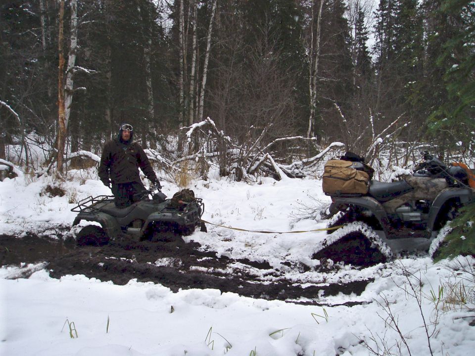 Guy on ATV stuck in mud, another ATV is being pulled out of the mud by a tow strap in a snowy forest