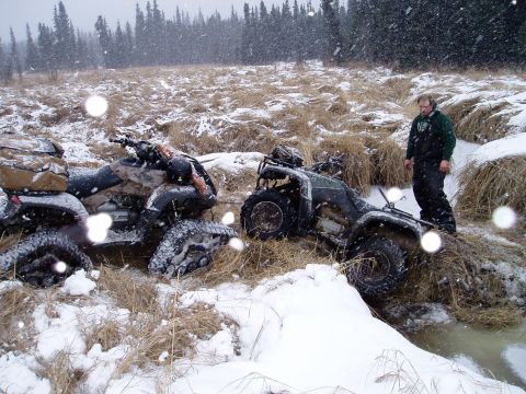 2000 Ford E350 - Two ATVs are stuck in a snowy, muddy ditch while a man watches.