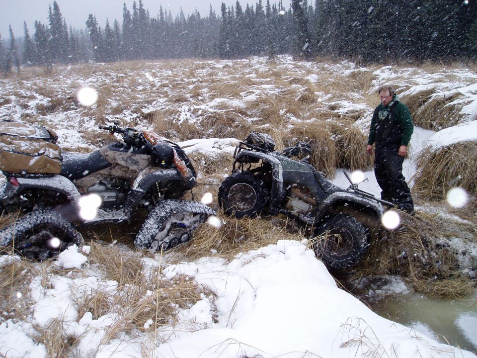 Two ATVs partially submerged in snow and mud, with a man standing to the right. Tall trees are in the background