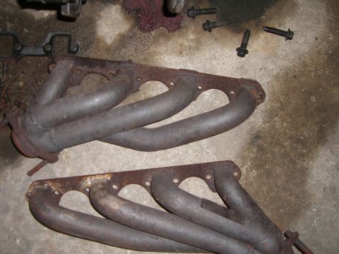 1988 Ford Bronco - Two rusty exhaust manifolds are shown with associated bolts and shop rags on a concrete surface.