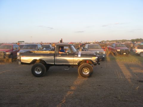 1979 Ford Bronco - A lifted pickup truck is parked in a dirt lot with several other vehicles and people visible in the background.