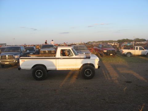1979 Ford Bronco - A lifted white pickup truck is parked in a field with other vehicles and people around.