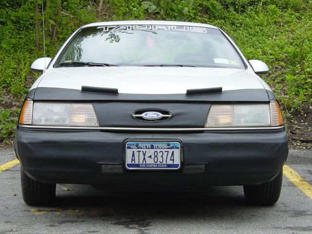 Front view of a white Ford Taurus with a black bra and New York plates