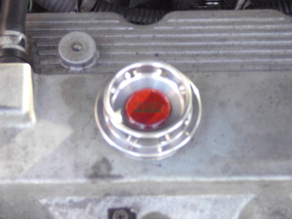 Engine oil cap with red insert and silver trim on a gray engine block