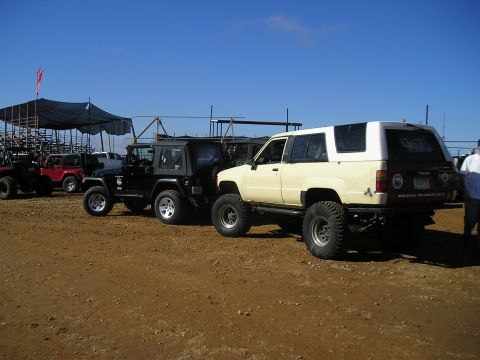 1989 Ford Bronco - Two modified SUVs, a Jeep and a Toyota, are parked side-by-side on a dirt lot under a clear blue sky.
