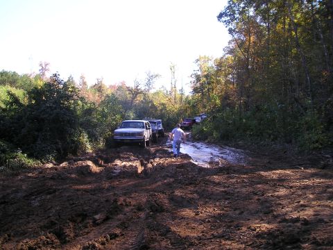 1989 Ford Bronco - A group of vehicles navigates a challenging muddy path surrounded by trees, with a person scouting ahead.