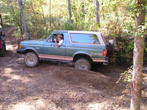 1989 Ford Bronco - A green 1989 Ford Bronco is shown off-roading on a dirt path, with another vehicle visible in the background.