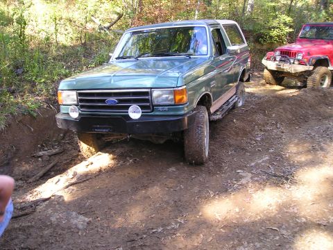 1989 Ford Bronco - Two SUVs navigating a muddy off-road path in a wooded area.