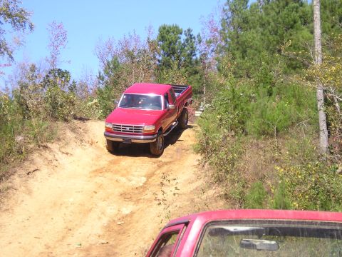 1989 Ford Bronco - A red pickup truck navigates an off-road trail with trees and brush surrounding the path.