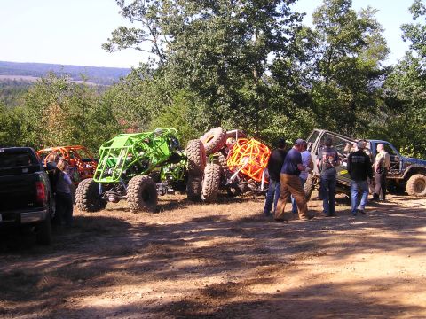 1989 Ford Bronco - A group of people stand around custom off-road vehicles and a pickup truck in a wooded area.
