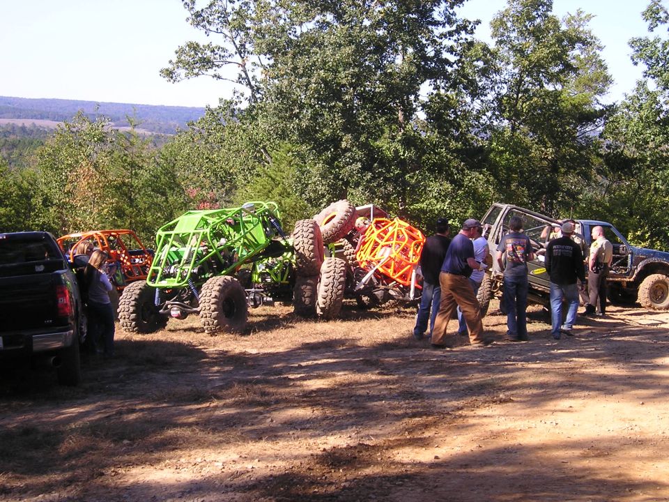 Several custom off-road buggies and a dark pickup truck parked on a dirt lot with trees in the background
