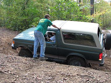 1989 Ford Bronco - A man in a green shirt steadies a Ford Bronco as it navigates a dirt path in a wooded area.