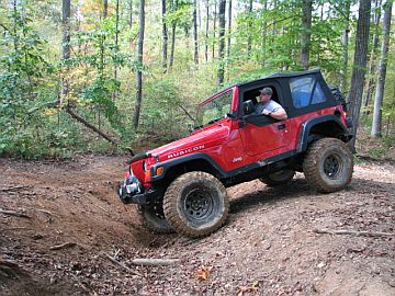 1989 Ford Bronco - A red Jeep Wrangler Rubicon navigates a challenging off-road course in a wooded area.