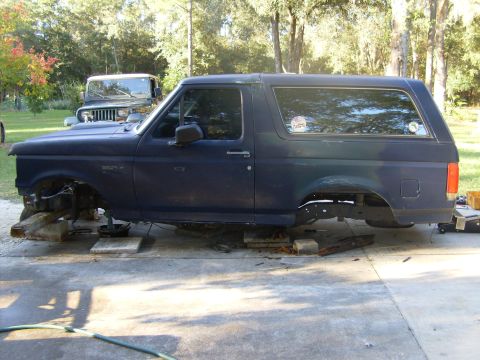 1995 Ford Bronco - Ford Bronco body is elevated on wooden blocks, front axle removed. A Jeep is parked behind it.