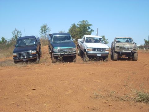 1992 Ford Bronco - Four pickup trucks are parked on a dirt incline under a clear blue sky.