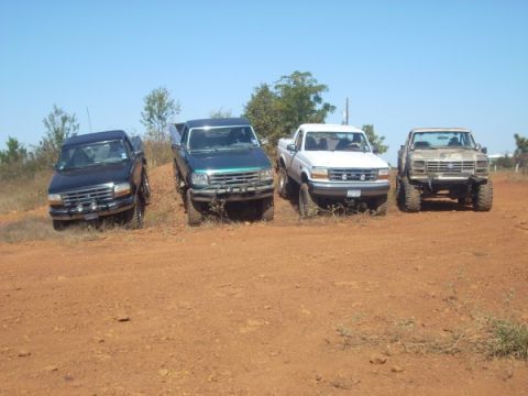 1992 Ford Bronco - A group of four pickup trucks, including a 1992 Ford Bronco, are parked on a dirt incline on a sunny day.