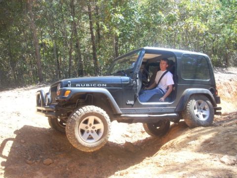 1992 Ford Bronco - A person drives a black Jeep Wrangler Rubicon off-road on a dirt trail with trees in the background.