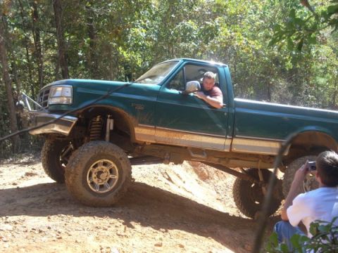 1992 Ford Bronco - A man drives a lifted green pickup truck on a wooded trail while another person captures the moment with a camera.