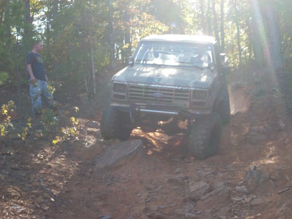 Ford Bronco with large tires navigating a muddy trail, person standing nearby