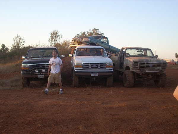 Man standing next to a black truck, with a white truck and a green truck behind it. A fourth, older truck is on the right