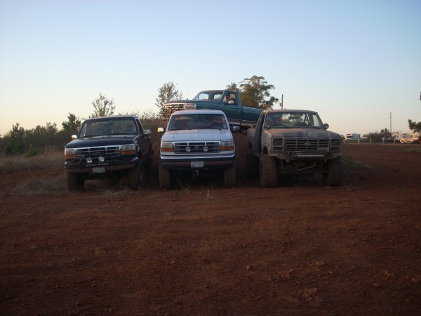 Four trucks, including a dark blue Ford, a white Ford, and a rusty older model, are parked on a dirt mound