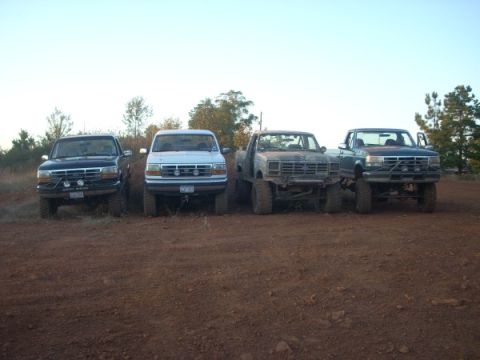 1992 Ford Bronco - Four trucks, including two Ford Broncos, are parked in a line on a dirt lot under a clear sky.