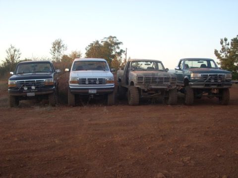 1992 Ford Bronco - Four lifted trucks, including a 1992 Ford Bronco, are parked side-by-side on a dirt lot.