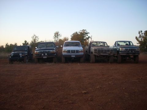 1992 Ford Bronco - A group of five modified trucks and SUVs are lined up on a dirt lot under a clear sky.