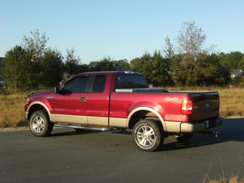 2005 Ford F-150 - A red pickup truck is parked on the side of a road with trees and houses in the background.