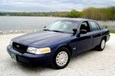 2003 Ford Crown Victoria - A dark blue 2003 Ford Crown Victoria P71 is parked near a waterfront with trees in the background.