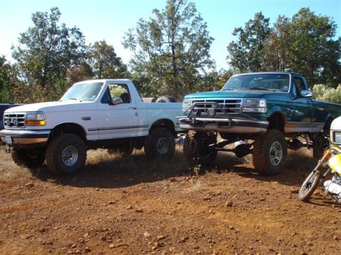 2008 International School Bus - Two lifted Ford F-150 pickup trucks are parked on a dirt lot under a clear sky.