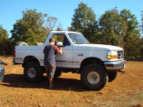 2008 International School Bus - A man stands next to a lifted white Ford Bronco on a dirt road surrounded by trees.
