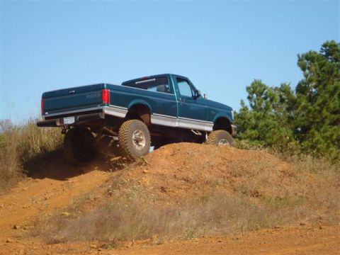 2008 International School Bus - A teal Ford pickup truck with a lifted suspension navigates a dirt hill under a clear blue sky.