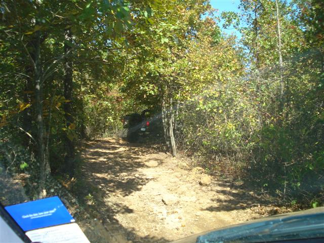 Dark off-road vehicle on a dirt path through trees, seen from inside another vehicle