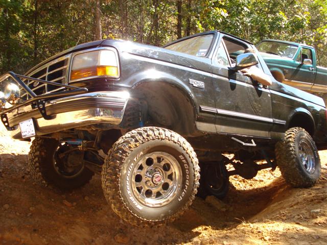 Black Ford Bronco with off-road tires on a dirt trail, another green truck behind it