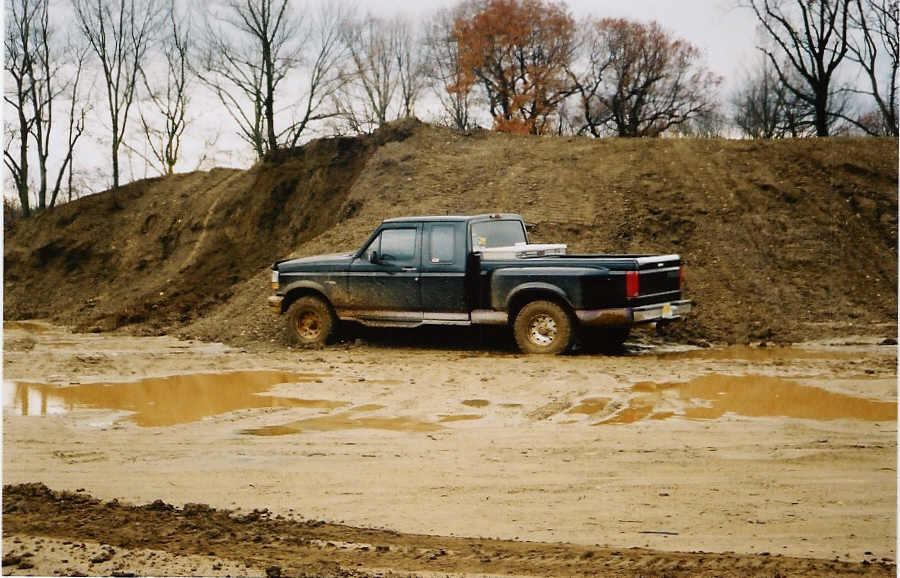 Dark green Ford extended-cab pickup parked in muddy lot with bare trees behind