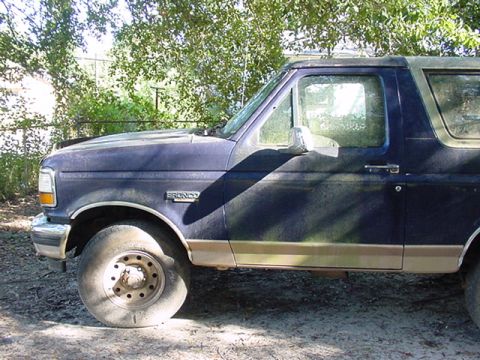 1994 Ford Bronco - A blue 1994 Ford Bronco is seen from the side, showing its tan trim and a flat tire.