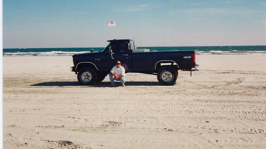Lifted dark blue square-body pickup parked on beach sand, man kneeling beside it