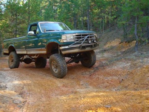 2008 International School Bus - A modified green pickup truck navigates a wooded trail with significant suspension lift.