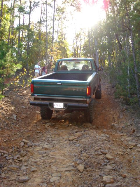 2008 International School Bus - A green pickup truck navigates a rocky, wooded trail under bright sunlight.