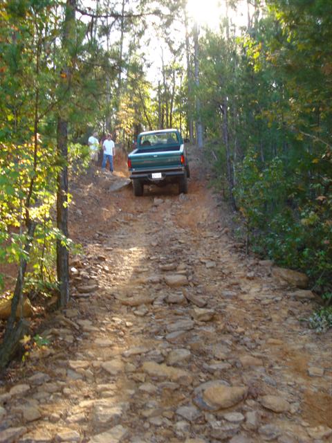 2008 International School Bus - A green pickup truck navigates a challenging, rocky trail through a wooded area.