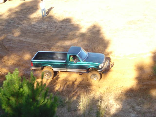 Green pickup truck with lifted suspension driving on a dirt track. A person stands in the background